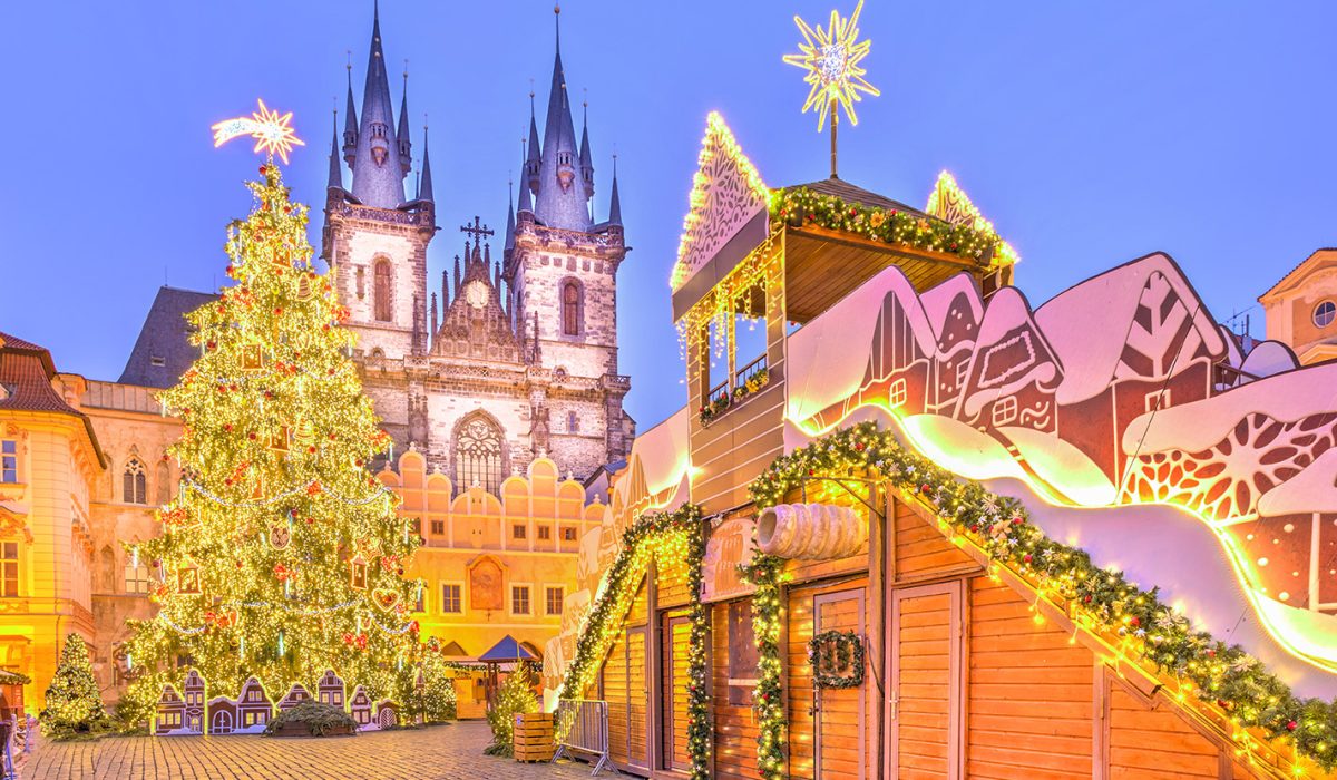 Świąteczny Jarmark Bożonarodzeniowy na Rynku Staromiejskim w Pradze, Czechy. Scena o zmierzchu lub w nocy, z dominującą, bogato oświetloną choinką i charakterystycznym, gotyckim Kościołem Marii Panny przed Tynem w tle. Na pierwszym planie widoczne są dekoracyjne drewniane stoiska ozdobione lampkami i motywami zimowymi, w tym domek z gwiazdą betlejemską na dachu.