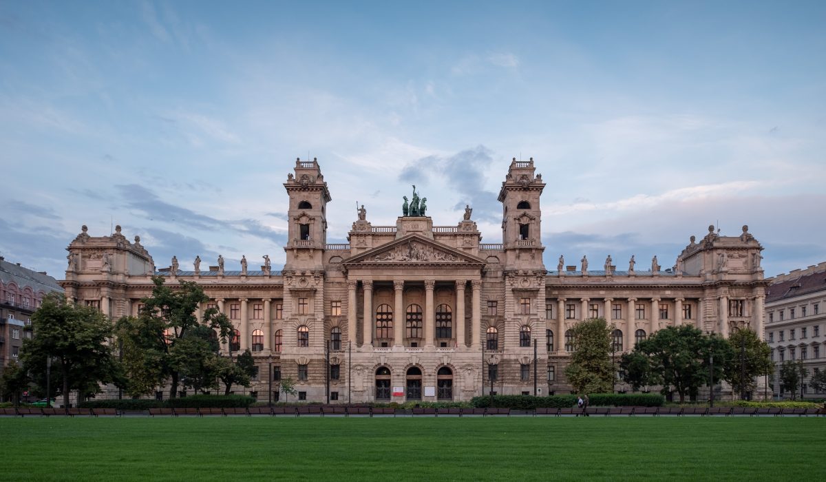 Frontalny widok na Muzeum Etnograficzne (Néprajzi Múzeum) w Budapeszcie, Węgry, budynek w stylu neorenesansowym z dwoma wieżami, kolumnowym portykiem i rzeźbami na dachu. Budynek jest widoczny z perspektywy obszernego, zielonego trawnika, który zajmuje pierwszy plan. Zdjęcie wykonane jest o zmierzchu, przy lekko zachmurzonym, jasnym niebie.