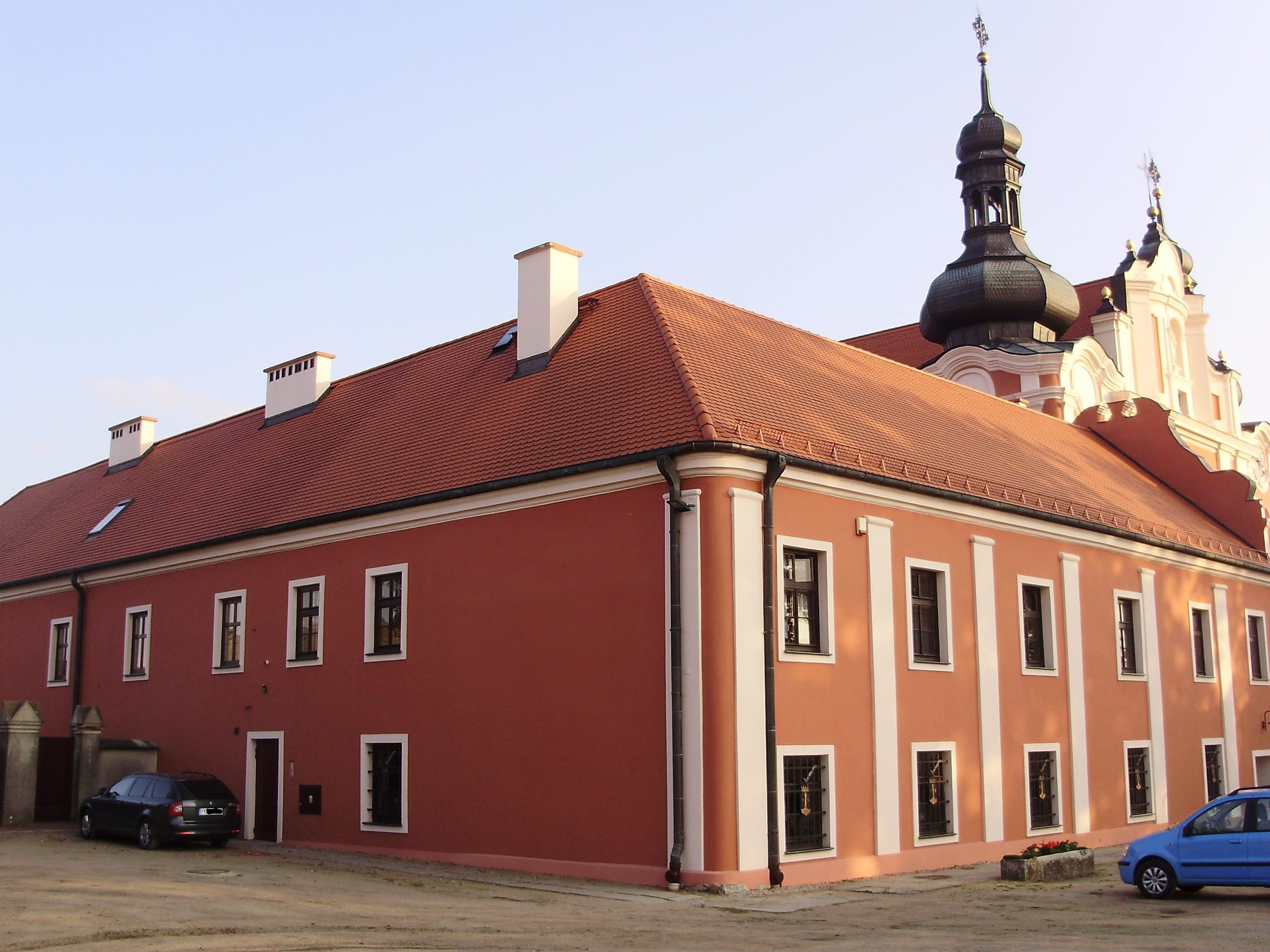Boczna fasada zabytkowego budynku sakralnego lub klasztoru o wyraźnym, ceglastoczerwonym kolorze i spadzistym dachu krytym czerwoną dachówką. Budynek ma symetrycznie rozmieszczone okna z ciemnymi ramami i jest ozdobiony białymi opaskami narożnymi. W prawym górnym rogu widoczna jest barokowa wieża kościelna (lub klasztorna) z czarnym hełmem. Na dole widać zaparkowane dwa samochody.