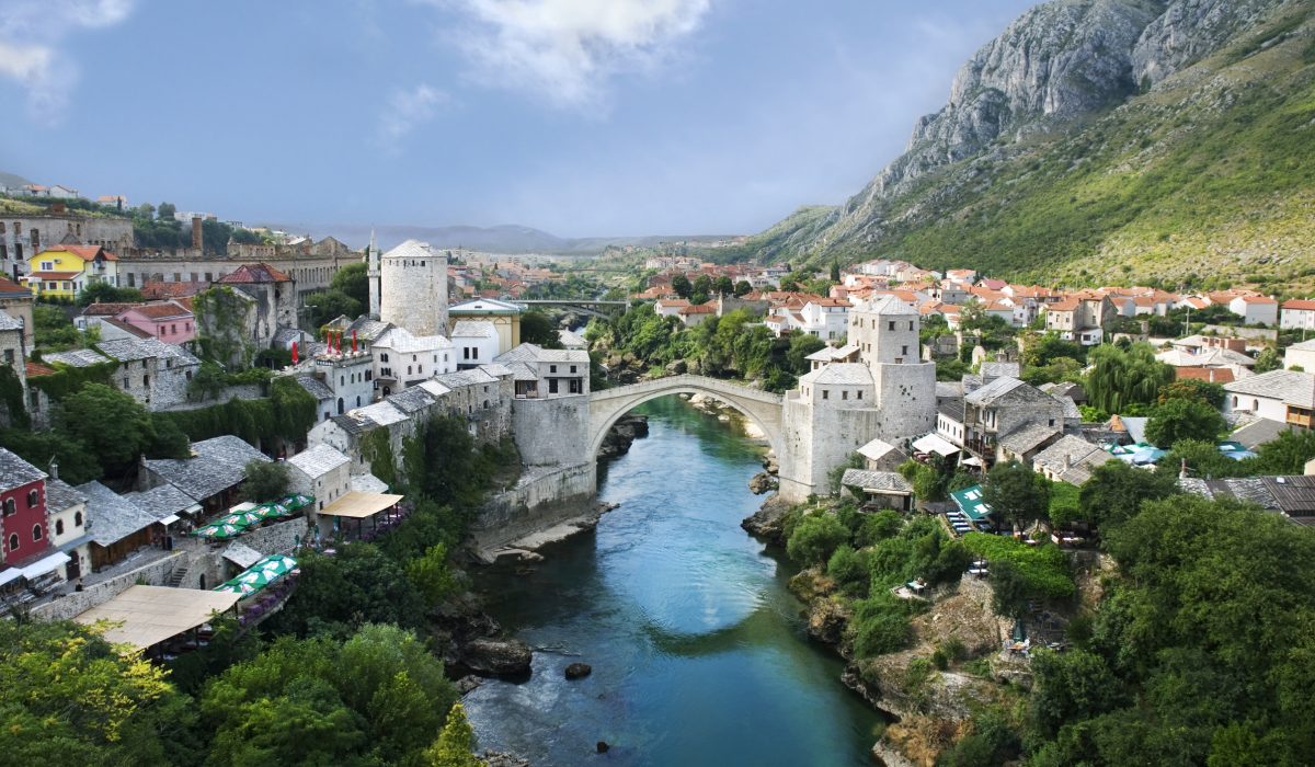 Szeroki widok na Stary Most (Stari Most) w Mostarze w Bośni i Hercegowinie, z renesansowym łukiem przerzuconym nad rzeką Neretwa. Po obu stronach mostu widać zabytkowe kamienne wieże (strażnice) i starówkę z osmańskimi domami o jasnych ścianach i szarych dachach. W tle rozciąga się dolina i strome, zielone zbocza gór.