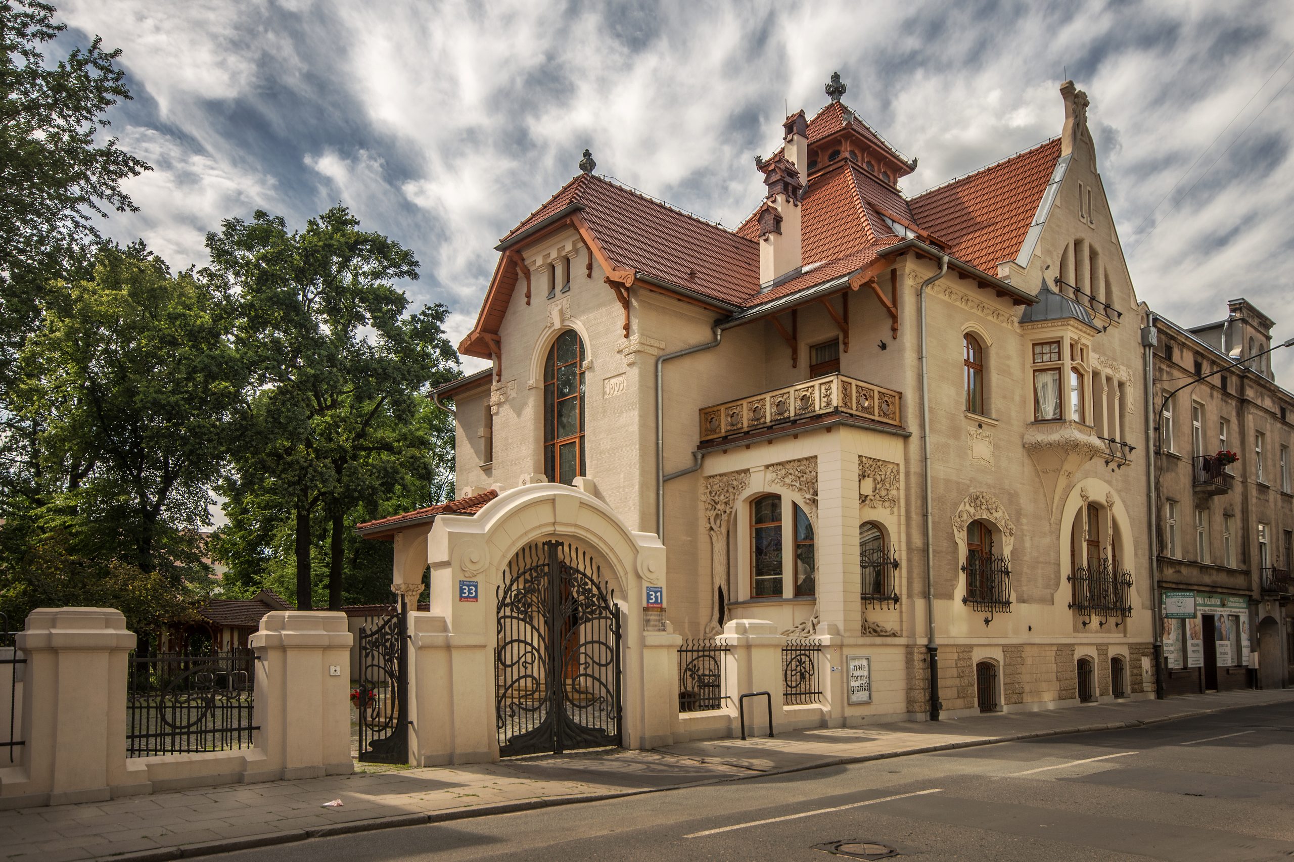 Willa Leopolda Kindermanna w Łodzi, przykład architektury secesyjnej, z jasną elewacją i czerwonym dachem. Budynek charakteryzuje się asymetryczną fasadą, wykuszami i bogato zdobioną bramą wjazdową z kutego żelaza, otoczoną kremowym murem. Po lewej stronie wysokie drzewa, po prawej ulica i sąsiednie kamienice. Dramatyczne niebo z puszystymi chmurami.