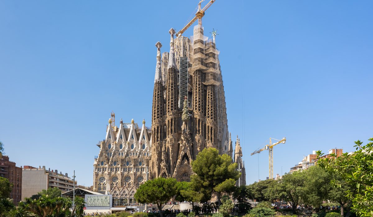Bazylika Sagrada Família w Barcelonie, Hiszpania, sfotografowana w słoneczny dzień od strony fasady Męki Pańskiej, która jest częściowo pokryta rusztowaniami w trakcie trwających prac budowlanych. Na pierwszym planie znajduje się park z niewielkim, zielonym stawem, oddzielającym świątynię od budynków mieszkalnych. Widoczne są dwa żółte żurawie budowlane.