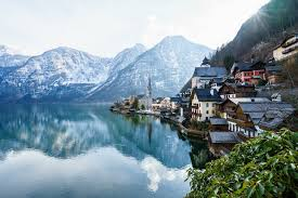 Pocztówkowy widok na miasteczko Hallstatt w Austrii, położone nad krystalicznie czystym jeziorem Hallstätter See. Widać charakterystyczne drewniane domy wciśnięte między zbocze góry a wodę, z wieżą kościoła pośrodku. W tle majestatyczne, częściowo ośnieżone Alpy odbijają się w tafli jeziora.