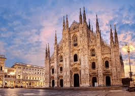 Katedra Duomo di Milano w Mediolanie, Włochy, sfotografowana o zmierzchu. Imponująca gotycka fasada jest oświetlona, a niebo ma odcienie różu i pomarańczu. Widoczna jest także część placu przed katedrą oraz fragment sąsiedniego budynku po lewej stronie.