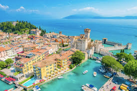Panoramiczne ujęcie malowniczego miasteczka Sirmione nad jeziorem Garda we Włoszech. Widać średniowieczny zamek Scaligero, otoczony turkusową wodą i połączony z lądem mostem, oraz gęstą zabudowę z kolorowymi dachami. W tle rozciąga się błękit jeziora i zarys gór.