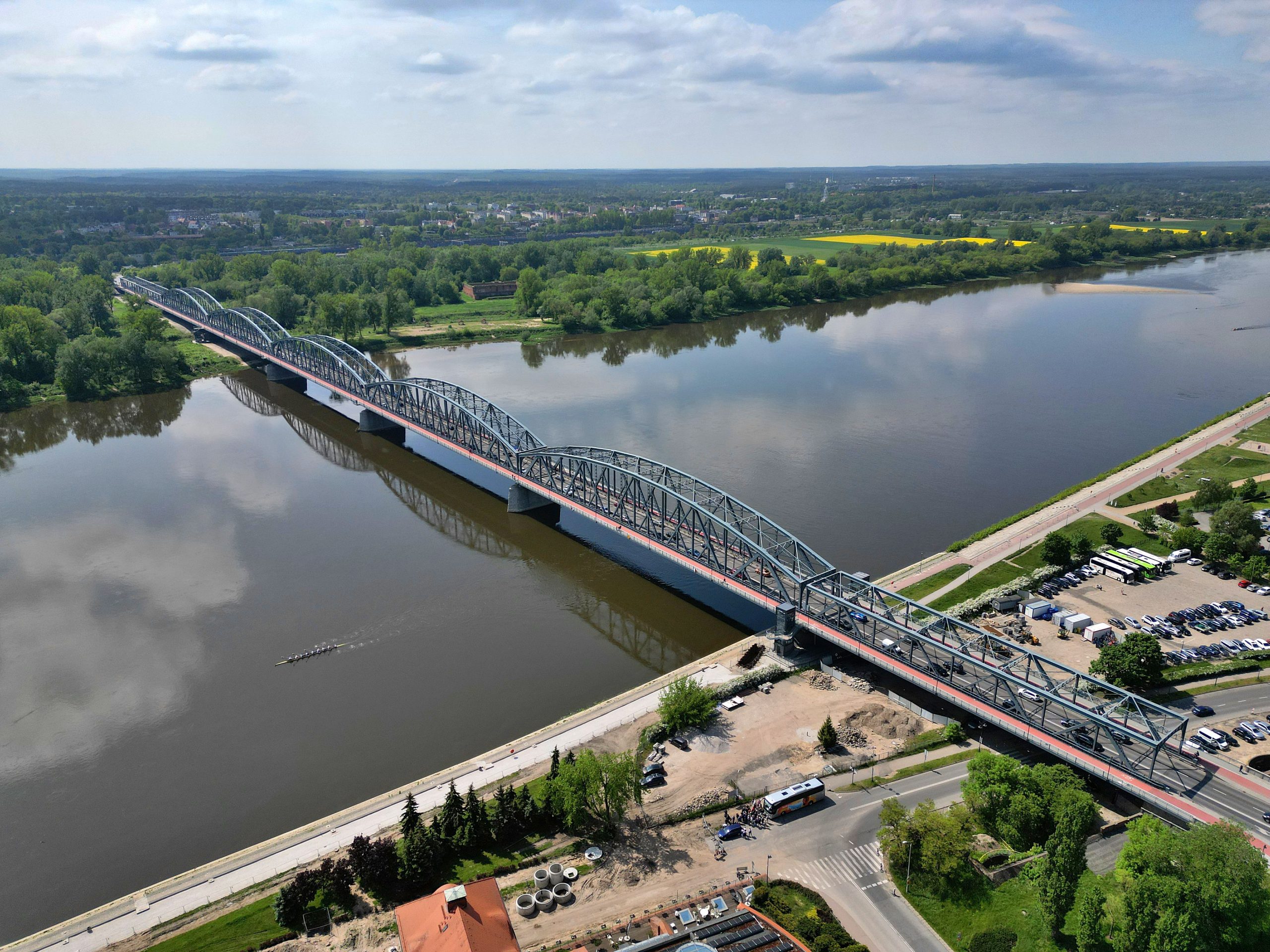 Ujęcie z drona przedstawiające długi, zabytkowy most kolejowo-drogowy im. Marszałka Józefa Piłsudskiego w Tczewie, rozciągający się nad szeroką, brunatną rzeką Wisłą. Most ma charakterystyczną kratownicową, stalową konstrukcję. W tle widać tereny zielone, pola uprawne, oraz zabudowania miasta. Na brzegu znajduje się parking i droga.