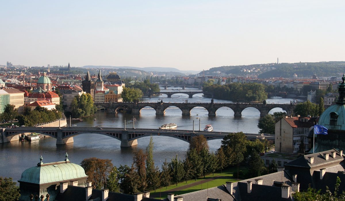 Szeroka panorama Pragi, Czechy, z widokiem na rzekę Wełtawę i serię historycznych mostów. Centralnie położony jest Most Karola z charakterystycznymi łukami i wieżami, a w tle widać inne mosty, zielone wzgórza, kopuły budynków i gęstą miejską zabudowę o czerwonych i zielonych dachach.