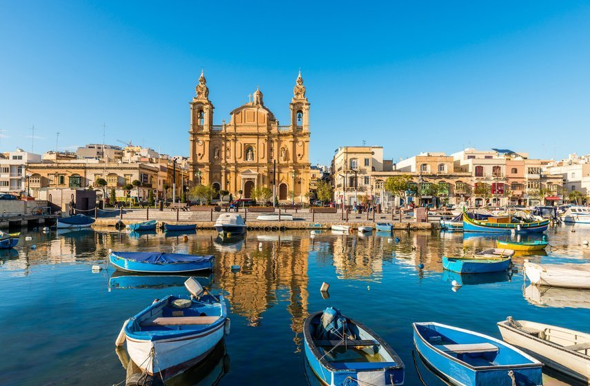Panoramiczny widok na port w Marsaxlokk na Malcie w słoneczny dzień. Na pierwszym planie na spokojnej wodzie cumują tradycyjne maltańskie łodzie rybackie, Luzzu, z których niektóre mają charakterystyczne jaskrawe kolory. W tle dominuje fasada kościoła Matki Bożej z Pompejów (Our Lady of Pompei) z dwiema dzwonnicami, otoczona budynkami mieszkalnymi. Kościół i budynki odbijają się w wodzie.