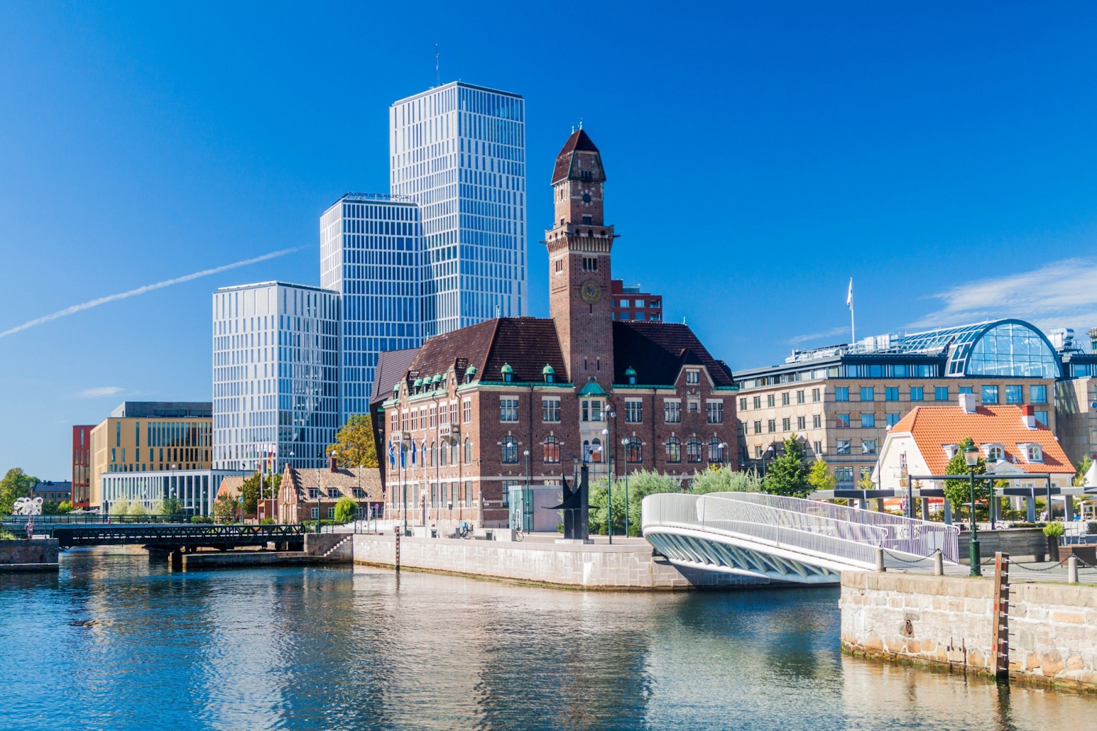 Panorama Malmö w Szwecji w słoneczny dzień. Na pierwszym planie widać ceglany budynek Starej Hali Targowej (Malmö Saluhall) z charakterystyczną wieżą zegarową, otoczony nowoczesnymi, przeszklonymi wieżowcami w tle. Na pierwszym planie przepływa kanał miejski, a po prawej stronie widać nowoczesny, biały most dla pieszych.