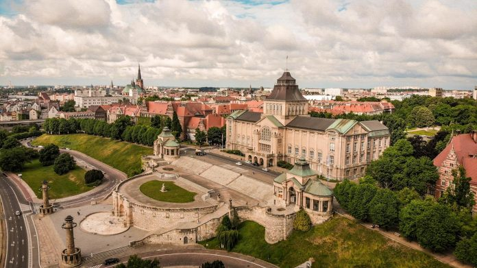 Lotniczy lub wysoki widok na Wały Chrobrego w Szczecinie, Polska, z widocznym budynkiem Muzeum Narodowego i tarasami schodzącymi w kierunku ulicy i terenów zielonych. W tle rozciąga się panorama Starego Miasta z charakterystycznymi wieżami i czerwoną ceglaną architekturą. Niebo jest pochmurne.