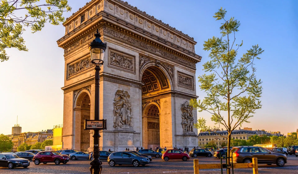 Łuk Triumfalny (Arc de Triomphe) w Paryżu, sfotografowany pod koniec dnia, gdy jego kamienna fasada jest oświetlona ciepłym, złocistym światłem zachodzącego słońca. Na pierwszym planie widoczna jest latarnia uliczna z tabliczką 'Place Charles de Gaulle', a wokół budowli parkują samochody. Po bokach ujęcia znajdują się gałęzie drzew.