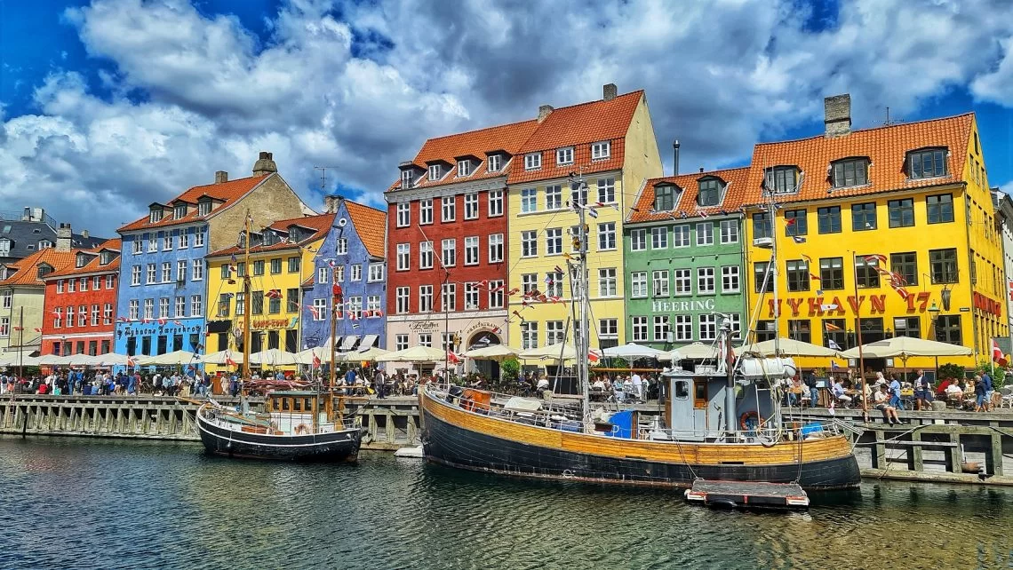 Słynna dzielnica Nyhavn w Kopenhadze, Dania, z jaskrawo kolorowymi, historycznymi kamienicami i żaglówkami (barkami) zacumowanymi w kanale. Nad wodą widać nabrzeże pełne turystów i stolików kawiarnianych. Niebo jest niebieskie z puszystymi, białymi chmurami.