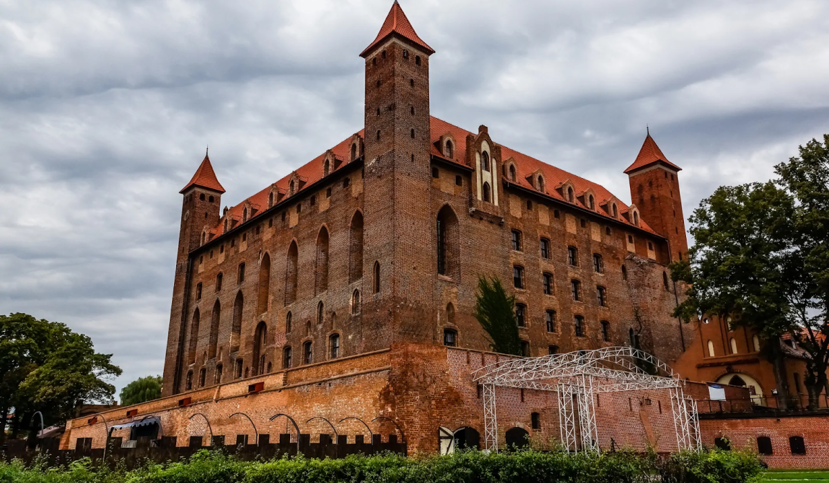 Zbliżenie na Wysoki Zamek w Malborku, z monumentalnym murem obronnym i czterema wysokimi wieżami narożnymi. Ciemna, ceglana fasada zamku kontrastuje z czerwonymi spadzistymi dachami. Na pierwszym planie widoczny jest niski żywopłot i nowoczesna, tymczasowa konstrukcja metalowa. Niebo jest pochmurne i dramatyczne.