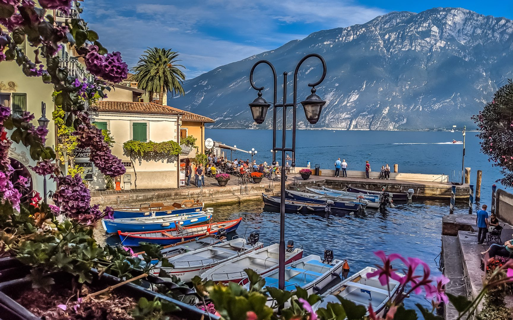Malownicza zatoczka portowa w Limone sul Garda we Włoszech. Na pierwszym planie widoczny jest tradycyjny czarny lampion i ramka z bujnej, purpurowej roślinności. W zatoce cumują kolorowe łodzie motorowe, a w tle widać kawiarnię nad wodą z gośćmi oraz strome, skaliste góry nad jeziorem Garda.
