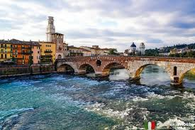 Średniowieczny rzymski most Ponte Pietra w Weronie we Włoszech, z szerokim ujęciem rzeki Adygi z jej bystrym, turkusowym prądem na pierwszym planie. Po lewej stronie widoczne są kolorowe kamienice i wieża, a w tle zarys historycznej zabudowy Włoch na tle wzgórz.