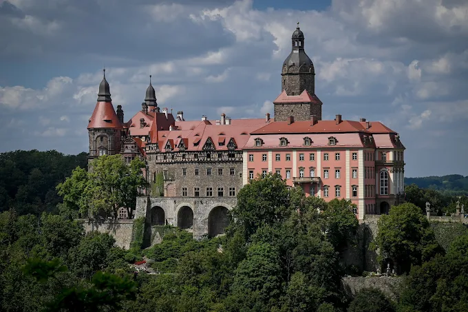 Majestatyczny Zamek Książ w Wałbrzychu (Polska), ujęty z daleka na tle pochmurnego, letniego nieba. Renesansowo-barokowy zamek stoi na zalesionym wzgórzu, eksponując ciemną, kamienną część środkową i jaśniejsze, różowo-kremowe skrzydła boczne z czerwonymi dachami. Wyróżniają się cztery wieże, w tym jedna centralna z charakterystycznym hełmem.